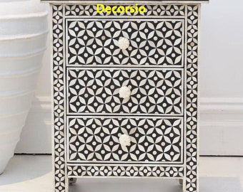 May include: A three-drawer nightstand with a black and white geometric pattern. The nightstand has a cream-colored frame and decorative knobs. The text "Decorcio" is visible on the top of the nightstand.