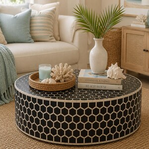 May include: A round coffee table with a black and white hexagonal pattern. On top sits a round woven tray containing a light blue candle and white coral, a white vase with green palm leaves, a white seashell, and two books titled Coastal Living and Beach House.