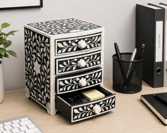 May include: A small black and white chest of drawers featuring four stacked drawers with floral inlay patterns and white flower-shaped knobs. The exterior surfaces are decorated with intricate leaf motifs, and the bottom drawer is pulled open.