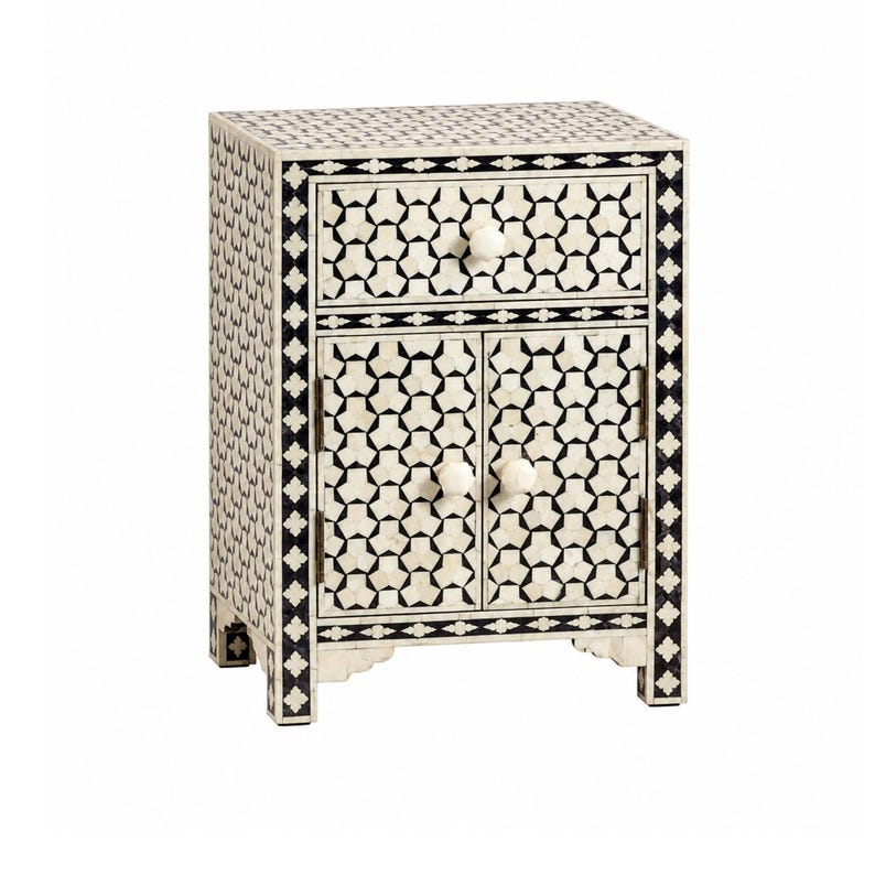 A rectangular bedside table featuring a black and white geometric inlay pattern. The piece includes one top drawer and two lower cabinet doors, each fitted with a small round white knob. The frame is decorated with a repeating floral border.