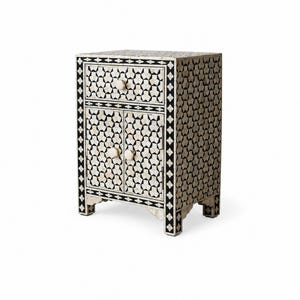 A rectangular bedside table featuring a black and white geometric inlay pattern. The piece includes one top drawer and two lower cabinet doors, each fitted with small, round, off-white knobs. The frame is accented with a black border.