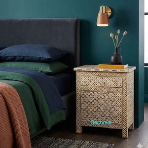 Könnte beinhalten: Beigefarbener Nachttisch mit geometrischem Muster, einer Schublade und einer Tür. Der Nachttisch steht neben einem Bett mit blauer und grüner Bettwäsche und einer orangefarbenen Decke. Eine schwarze Vase mit Trockenblumen steht darauf. Der Tisch ist ca. 60 cm hoch.