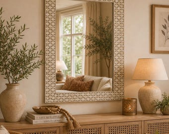 May include: A rectangular wall mirror featuring a frame with a repeating geometric pattern in beige and white. The mirror reflects a room interior with a window, a lamp, and a sofa with brown cushions, providing a clear view of the surroundings.