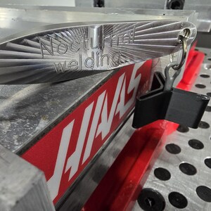 May include: A close-up of a metal object with the text "Nocturnal welding" engraved on it. The object has a unique design with spiral patterns and is attached to a black clip. The background includes a red and white sign.
