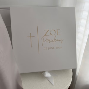 May include: A white gift box featuring gold text and a cross design. The box reads "ZOE Pericleous 02 JUNE 2024". It is tied with a white ribbon. The box is square and likely made of card.