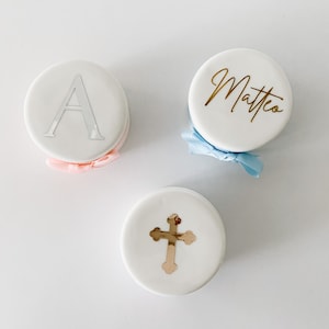 May include: Three white, round favours with decorative elements. One has a silver letter "A", another has the name "Matteo" in gold script, and the third features a gold cross. Each favour is tied with a ribbon in either pink or blue.