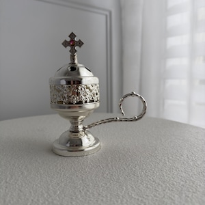 May include: A silver censer with a cross finial and a handle. The censer features a floral design around the body and small holes at the top. The handle is curved and textured. The cross has a small, colourful gem in the centre.