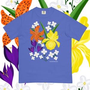 Puede incluir: Una camiseta azul con un diseño floral. El diseño incluye lirios tigrados naranjas, iris amarillos, crocus morados y flores blancas. La camiseta tiene un corte relajado. La firma del artista está en la parte inferior.