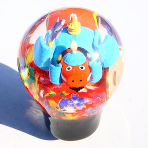 May include: A red, blue and yellow figurine of a cartoon character inside a clear glass sphere. The figurine is sitting on a red, yellow and blue patterned fabric.