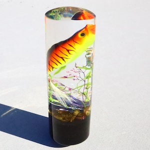 May include: A clear cylinder filled with resin containing a yellow, orange, and black fishing lure, green and pink plants, and a white feather. The cylinder has a black base.