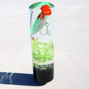May include: A clear cylinder filled with resin, containing a green, white, and red fishing lure with a hook. Green plant-like material is also embedded in the resin.