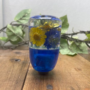 Puede incluir: Un pomo de palanca de cambios de vidrio transparente con resina azul y flores amarillas y azules prensadas.