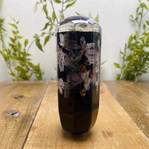 May include: A decorative, cylindrical object with a faceted top. The interior is dark, filled with dried flowers in shades of pink and white. The object is set on a wooden surface, with greenery in the background.