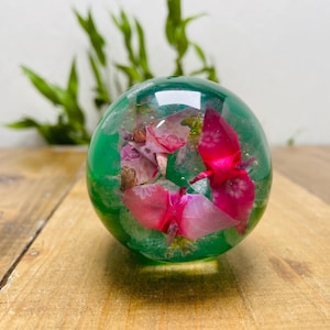 Puede incluir: Un pisapapeles esférico transparente con una base verde vibrante, que encierra elementos florales rosas y blancos. El pisapapeles se encuentra sobre una superficie de madera, con follaje verde en el fondo.