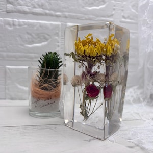 Può includere: Un blocco rettangolare in resina trasparente contenente girasoli gialli, piccoli fiori bianchi e boccioli di rosa rossa. Accanto si trova un piccolo portacandele in vetro con un cactus verde e la scritta Life with a Cactus.