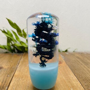 Puede incluir: Un objeto cilíndrico transparente con una base azul y una escultura de dragón negro en su interior. El dragón es detallado y está rodeado de detalles azules. La parte superior del objeto tiene un aspecto translúcido color ámbar.