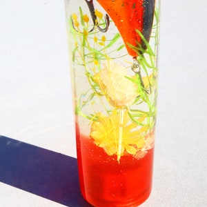 May include: A clear glass cylinder filled with a red liquid and a fishing lure. The lure is orange and black with a yellow and green flower inside the cylinder.