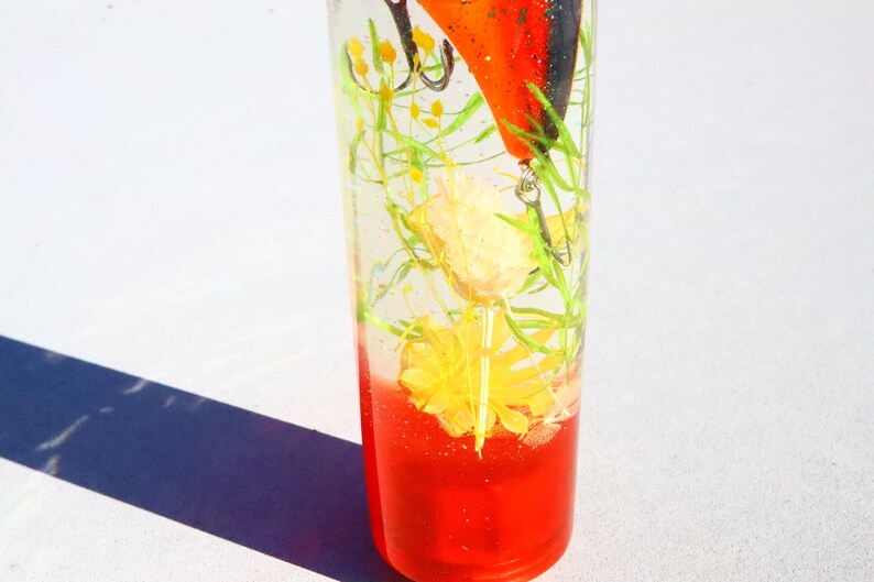 May include: A clear glass cylinder filled with a red liquid and a fishing lure. The lure is orange and black with a yellow and green flower inside the cylinder.