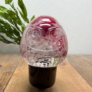 Può includere: Una statuetta Daruma in vetro trasparente con una parte superiore arrotondata e una solida base cilindrica nera. Il vetro presenta un volto inciso con grandi occhi circolari e una bocca sorridente, con il kanji giapponese per la fortuna, 福, inciso.