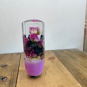 May include: A clear glass cylinder filled with dried pink and purple flowers, with a light purple base. The cylinder is sitting on a wooden surface.