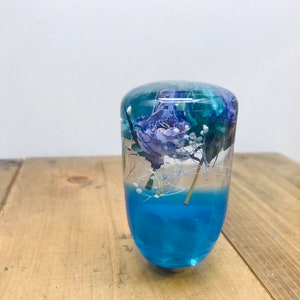 Könnte beinhalten: Ein transparenter Schaltknauf aus Harz mit blauer Basis und einer konservierten Blume im Inneren. Die Blume ist helllila mit weißen Akzenten.