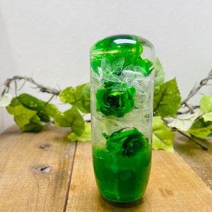 Könnte beinhalten: Ein transparenter Glaszylinder gefüllt mit konservierten grünen Blumen. Die Blumen sind in einem Strauß angeordnet und beinhalten Rosen und andere grüne Pflanzen. Der Zylinder steht auf einer Holzoberfläche.