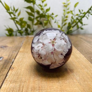 May include: A clear, spherical paperweight with white flower blossoms encased within a dark purple resin. The paperweight is resting on a wooden surface, with green foliage in the background.