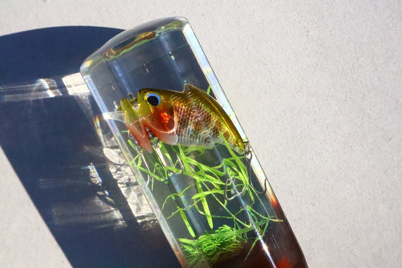 May include: A clear resin paperweight containing a gold and red fishing lure. The lure is surrounded by green plastic grass.