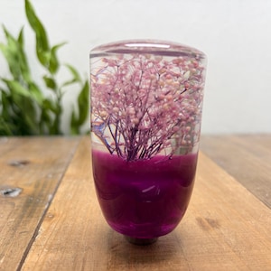 May include: A cylindrical gear shift knob made of clear resin, featuring a cluster of dried pink baby's breath flowers suspended in the upper section and a solid, deep purple opaque base at the bottom, creating a distinct two-tone appearance.