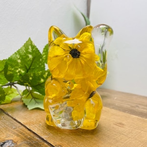 Op de afbeelding: Een geel glazen kattenbeeldje met een enkele gele bloem erin geperst. De kat zit rechtop met zijn rechterpoot omhoog, alsof hij zwaait.