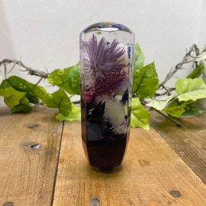 Op de afbeelding: Een transparante glazen versnellingspookknop met paarse en witte bloemen ingekapseld in hars. De knop heeft een donkerpaarse basis.