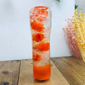 Puede incluir: Un jarrón de vidrio transparente en forma de cilindro lleno de flores naranjas y agua. El jarrón tiene una base naranja gruesa.