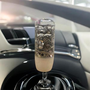 May include: A clear, cylindrical car gear shift knob filled with various silver-toned jewelry pieces, including chains, beads, and pearls. The bottom portion is a frosted white, and it sits atop a chrome base. The background shows a car interior.