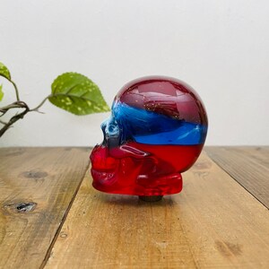 May include: Red, white and blue glass skull gear shift knob. The skull is made of clear glass with red, white and blue layers. The skull is sitting on a wooden surface.