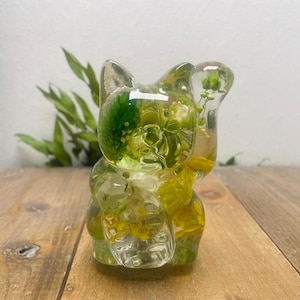 May include: A clear resin squirrel ornament with yellow and green flowers encased within. The squirrel is standing, holding a clear object. The ornament sits on a wooden surface, with green foliage visible in the background.