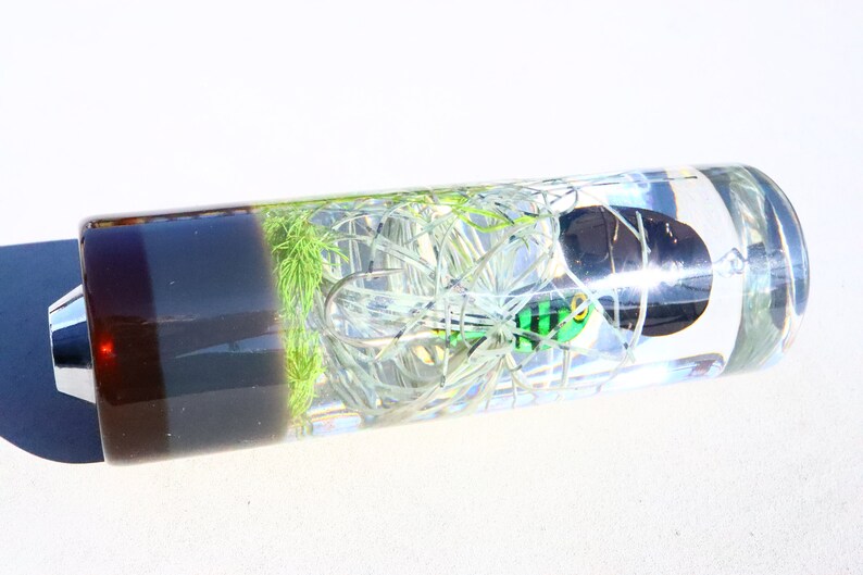 May include: A clear glass cylinder containing a green and black fishing lure. The lure is surrounded by white and green fishing line. The cylinder has a brown and grey base.