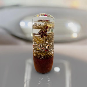 Puede incluir: Un objeto cilíndrico transparente de resina lleno de joyas doradas, piedras preciosas rojas y perlas. La parte inferior es un brillo marrón rojizo. Las joyas incluyen cadenas, anillos y un diseño floral. El objeto se muestra sobre una superficie reflectante.