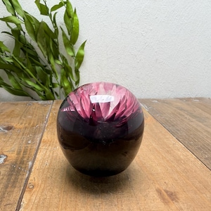 May include: A clear, spherical paperweight with a dark purple base and a vibrant pink floral design encased within. The paperweight sits on a rustic wooden surface, with green foliage in the background.