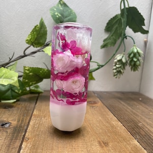 May include: A clear glass cylinder filled with pink and white flowers, preserved in resin. The cylinder has a white base and is approximately 12.5 cm tall.