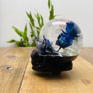 Puede incluir: Una calavera de resina transparente con base negra, que encierra flores azules. La calavera está sobre una superficie de madera, con follaje verde al fondo. La resina es transparente, permitiendo ver las flores.