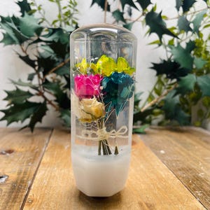 May include: A clear glass cylinder with a white base contains a bouquet of preserved flowers. The flowers are pink, yellow, green, and blue.