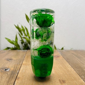 Puede incluir: Objeto decorativo rectangular transparente con tres rosas verdes. La base es de un verde vibrante. Colocado sobre una superficie de madera, con vegetación en el fondo. Aproximadamente 15 cm de altura.