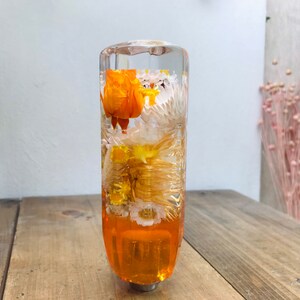 May include: A clear glass gear stick knob with dried flowers encased in resin. The flowers are white, yellow, and orange.