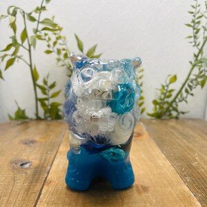 Peut inclure: Figurine décorative en forme de chien-lion, composée de résine transparente, blanche et bleue. La base et les détails sont bleu vif. L'objet est posé sur une surface en bois, avec des plantes vertes en arrière-plan.