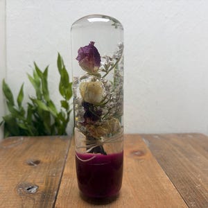 Puede incluir: Un pisapapeles cilíndrico de resina transparente que contiene flores secas, incluyendo una rosa morada y capullos blancos, suspendidos en el material. La base del cilindro es de un color morado oscuro sólido y opaco.