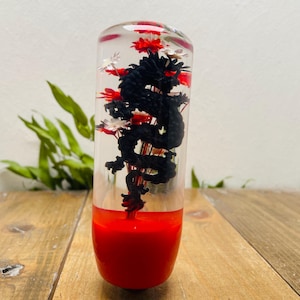 Puede incluir: Objeto decorativo cilíndrico transparente con base y parte superior rojas. En el interior, una figura de dragón negro está rodeada de elementos florales rojos y blancos. El objeto está sobre una superficie de madera.