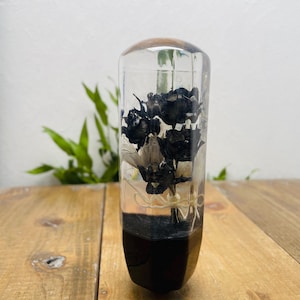 Op de afbeelding: Een helder, cilindrisch kunstwerk van hars met een donkere basis en een houten bovenkant. De hars omsluit zwart-witte gedroogde bloemen. Het stuk wordt op een houten oppervlak gepresenteerd.