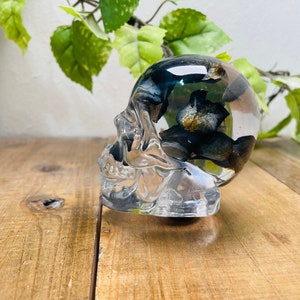 Peut inclure: Un crâne en verre transparent avec des accents floraux noirs et dorés à l'intérieur. Le crâne est posé sur une surface en bois.