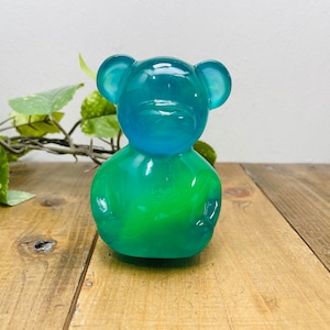 Könnte beinhalten: Eine durchscheinende Bärenfigur mit einem Farbverlauf von Blau- zu Grüntönen. Der Bär hat abgerundete Ohren und eine glatte, glänzende Oberfläche. Die Figur steht auf einer Holzoberfläche.
