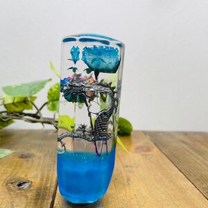 Peut inclure: Un objet rectangulaire transparent en résine avec une base bleue et une rose bleue enfermée à l'intérieur. Une figurine de dragon argenté est suspendue au milieu. Le dessus a un motif bleu. L'objet est sur une surface en bois.
